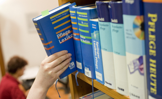 Eine Person greift aus einem Bibliotheksregal ein Fachbuch zum Thema Pflege